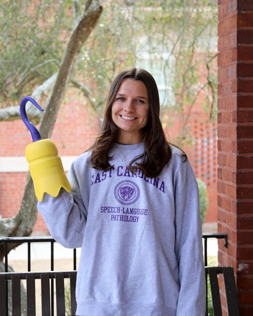 female student wearing gray sweatshirt with the words East Carolina Speech-Language Pathology and holding a 3D printed hook