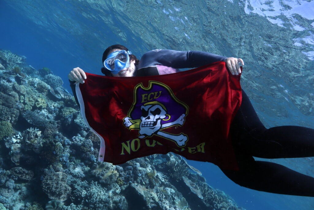 female student snorkeling and holding the ECU No Quarter flag