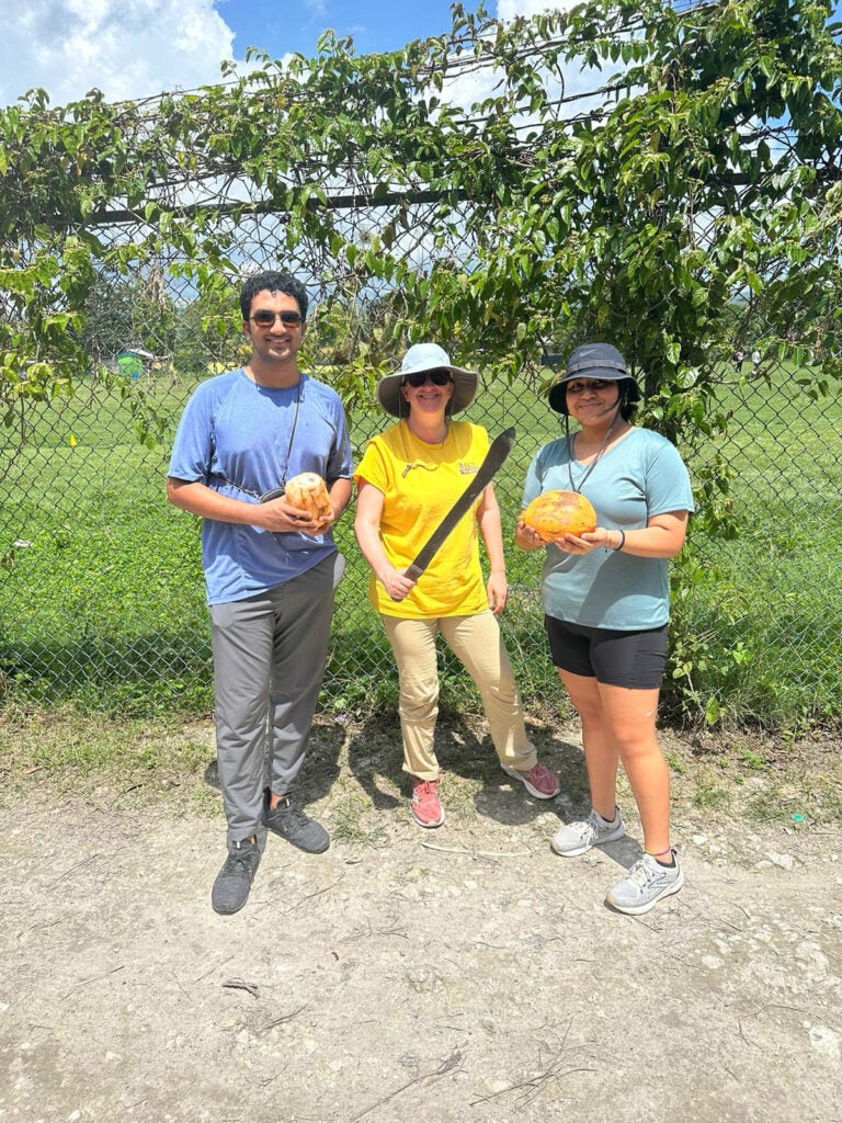 three people stand in front of a fence with two holding coconuts and one holding a machete